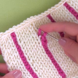Hand holding crochet hook to add vertical stripe in pink yarn with knitted swatch in stockinette stitch in white yarn color.