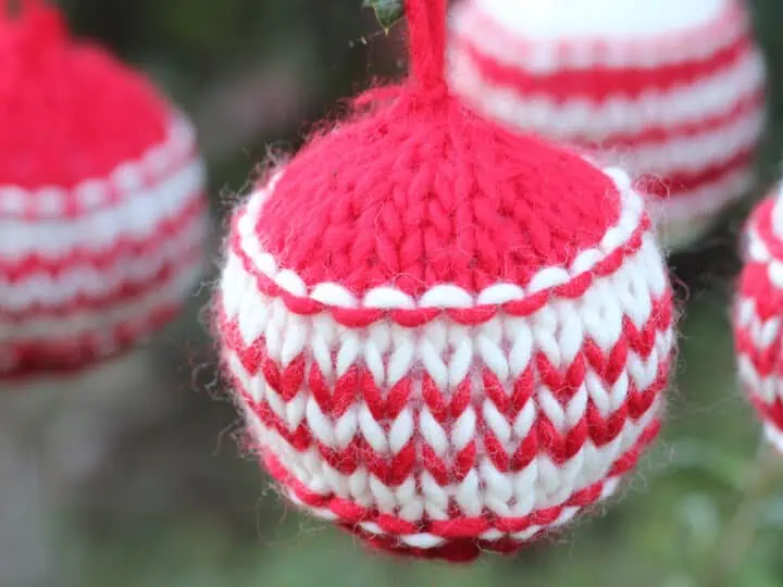 Knitted Christmas Ball Ornaments in red and white yarn colors hanging from outdoor greenery branches.