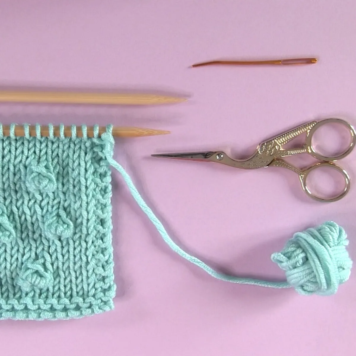 Knitting Materials with Bobble Stitch swatch in light blue yarn color, knitting needles, scissors, and a tapestry needle.