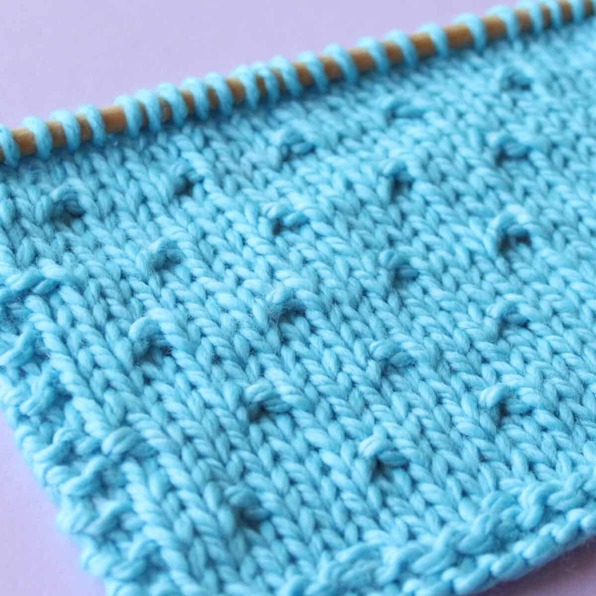 Swatch of Simple Seed Stitch Pattern in blue yarn on knitting needle atop a purple background.