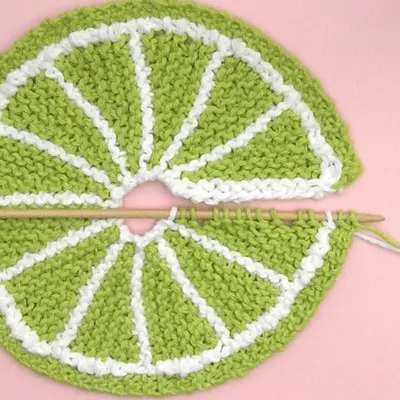 Knitted dishcloth in fruit slice design with green yarn on needle.