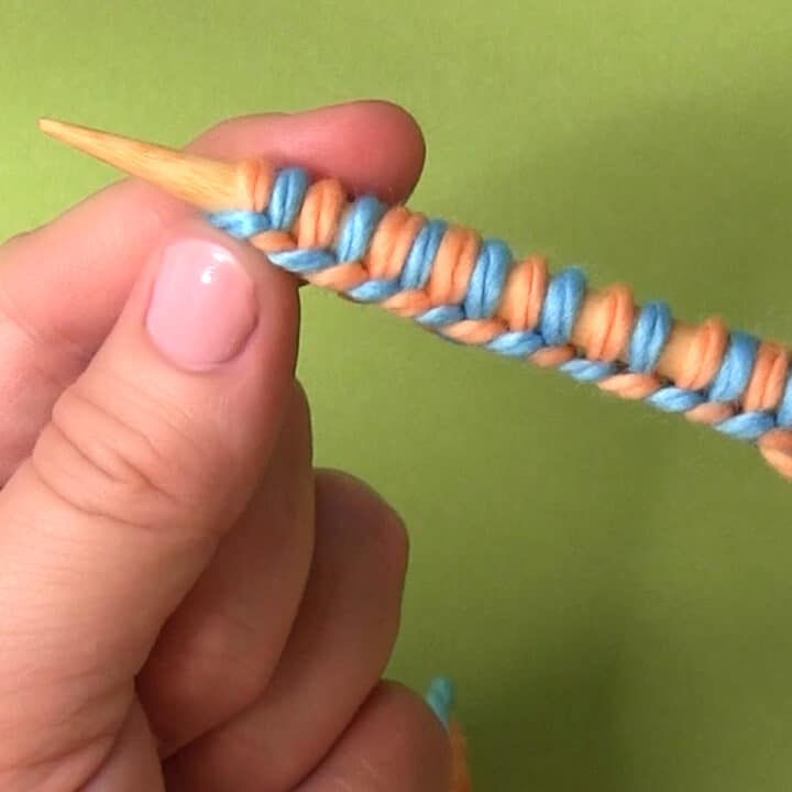 Hands holding knitting needle with two different yarn colors cast on.