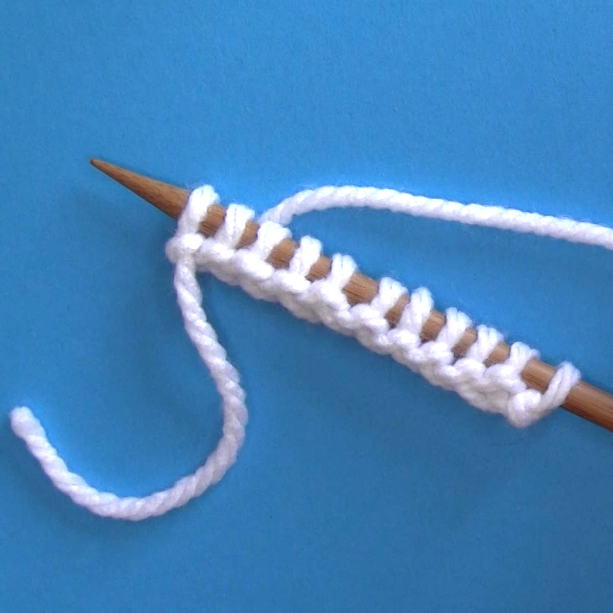 Knitting needle with stitches cast on purlwise in white color yarn on a blue background.