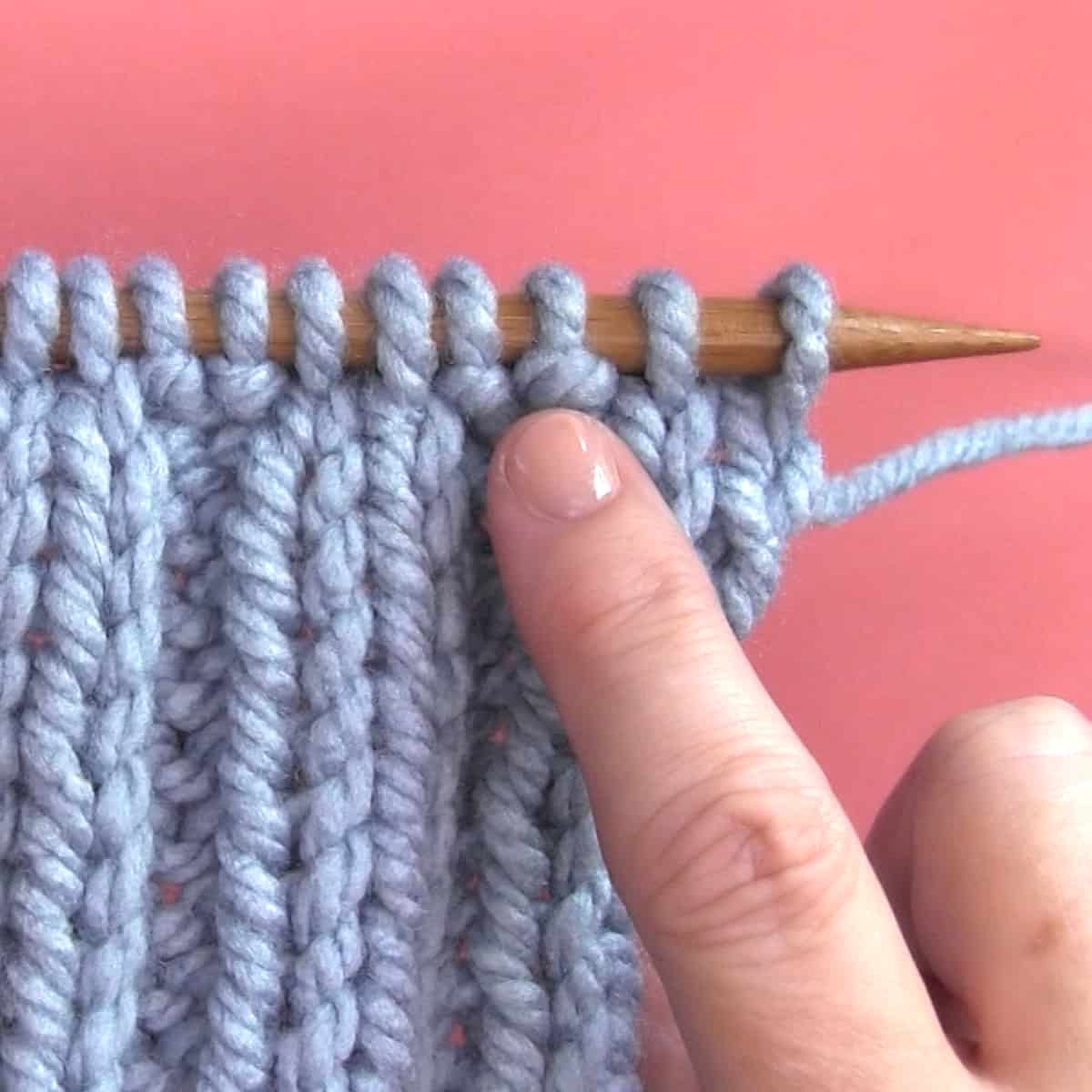 Finger pointing out a purl stitch on a knitting needle with yarn.