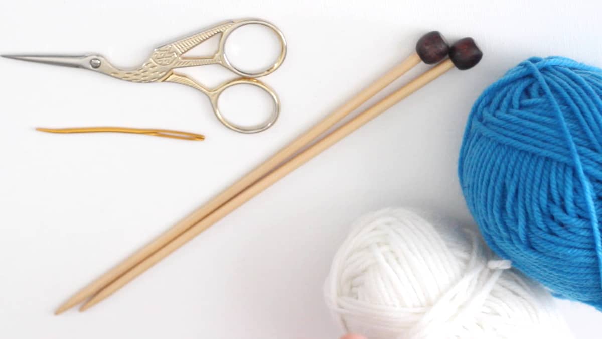 Tools and materials to knit a coffee cozy with scissors, knitting needles, yarn, and a tapestry needle.