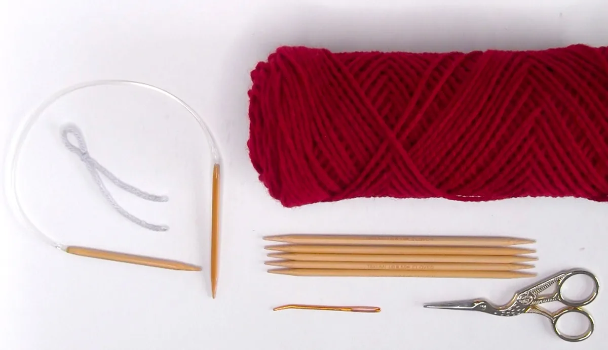 Knitting supplies of burgundy worsted yarn, knitting needles, scissors, and tapestry needle.
