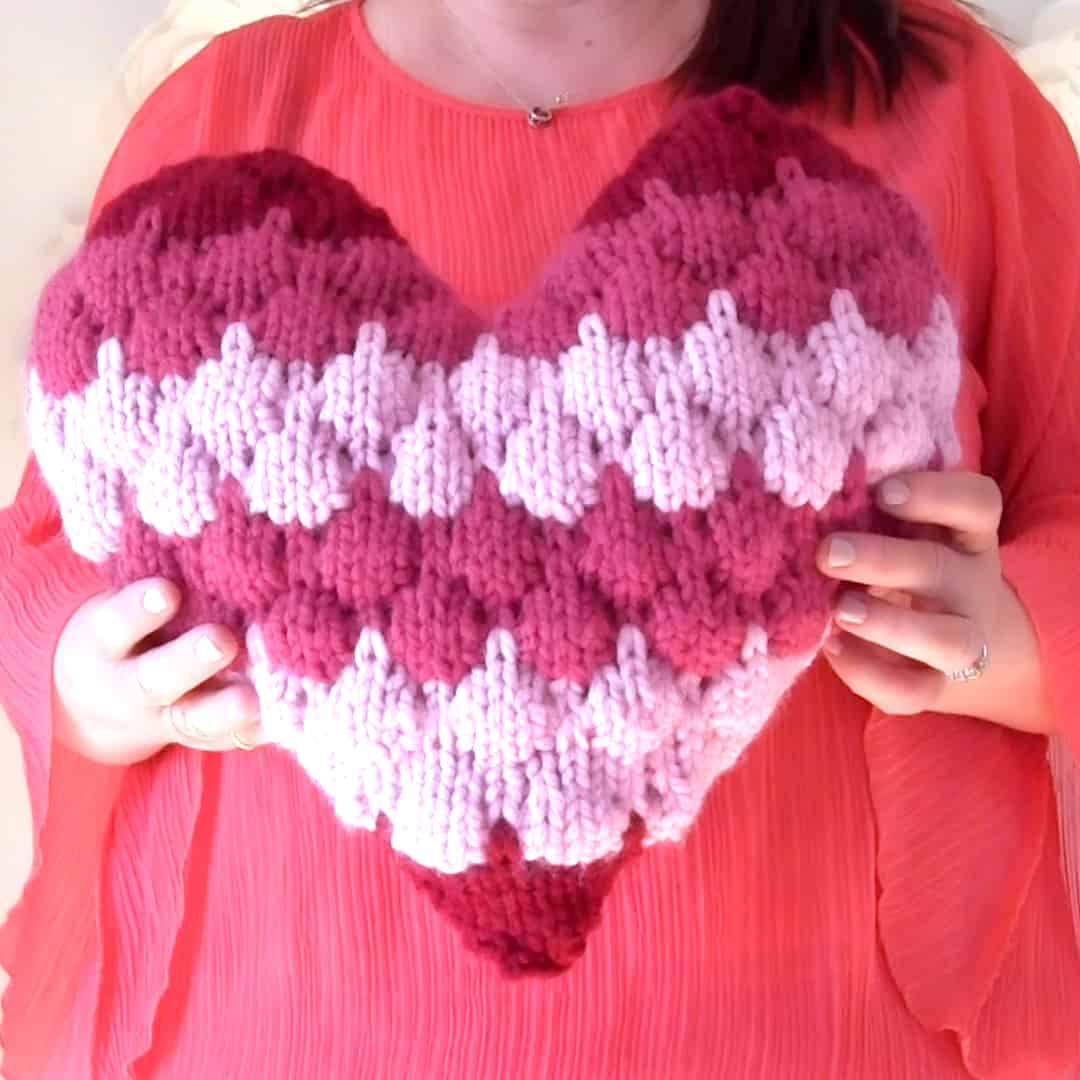 Woman holding heart shaped knitted pillow in bubble stitch.