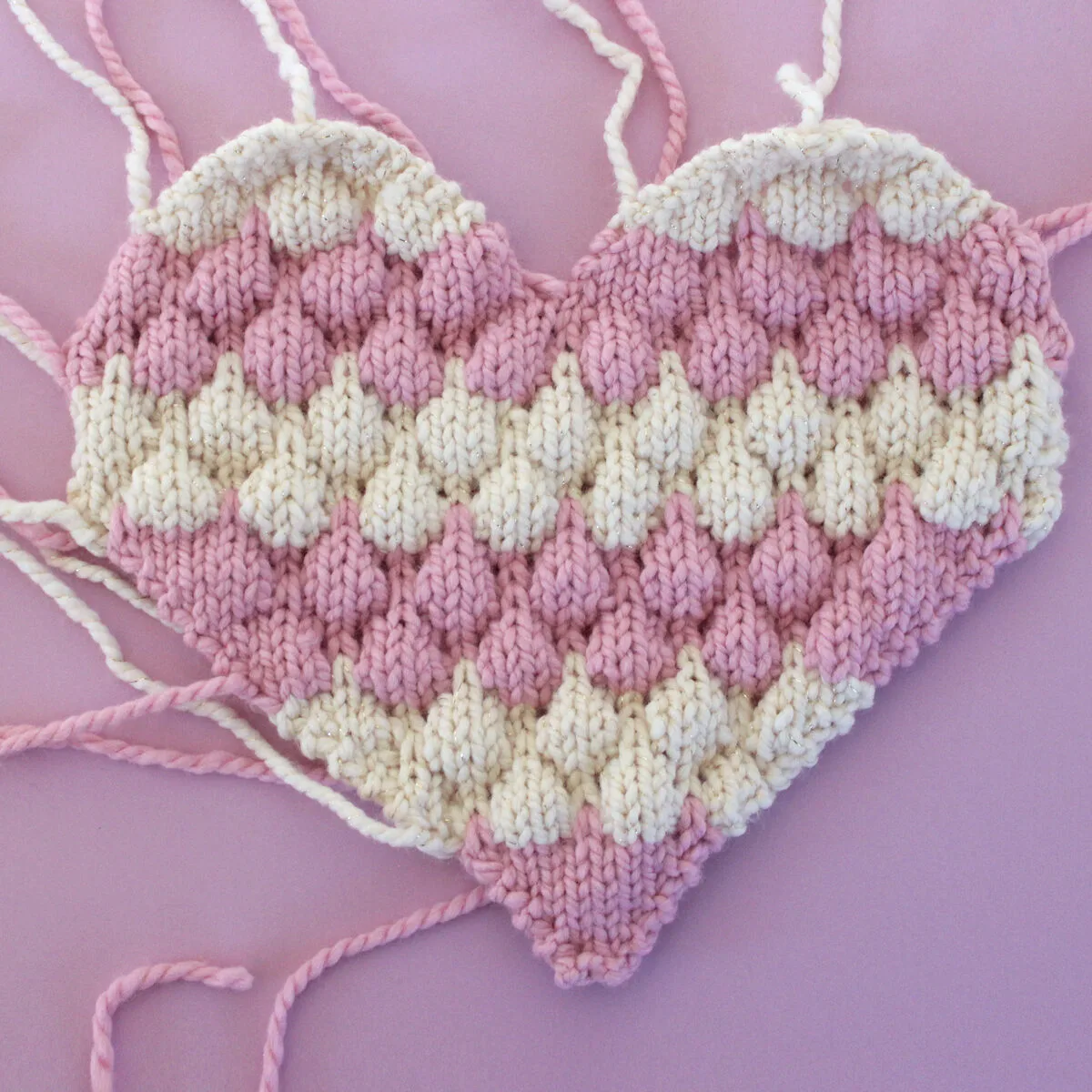 One side of the heart pillow in bubble stitch in pink and white yarn colors.