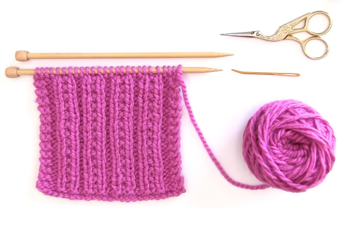 Knitting supplies of hot pink yarn color, knitting needles, scissors, and a tapestry needle.