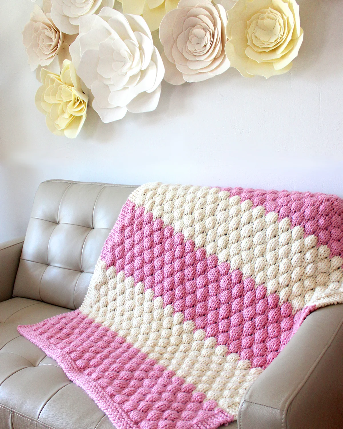 Knitted Bubble Stitch Blanket in pink and white yarn colors on a couch and felt flowers on the wall.