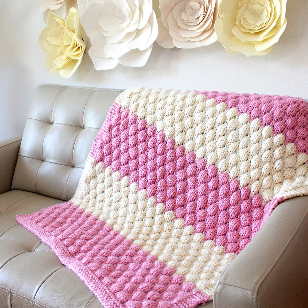 Knitted Bubble Stitch Blanket in pink and white yarn colors on a couch and felt flowers on the wall.