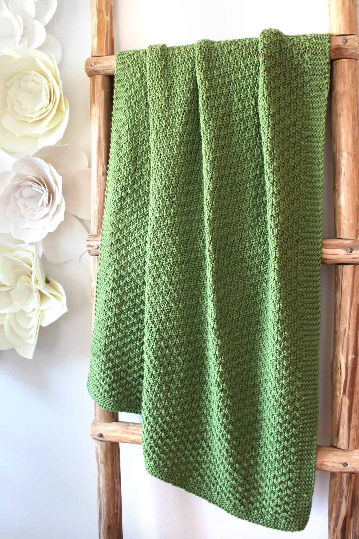 Knitted Double Moss Stitch draped on Blanket Ladder in green yarn color with felt flowers on wall.