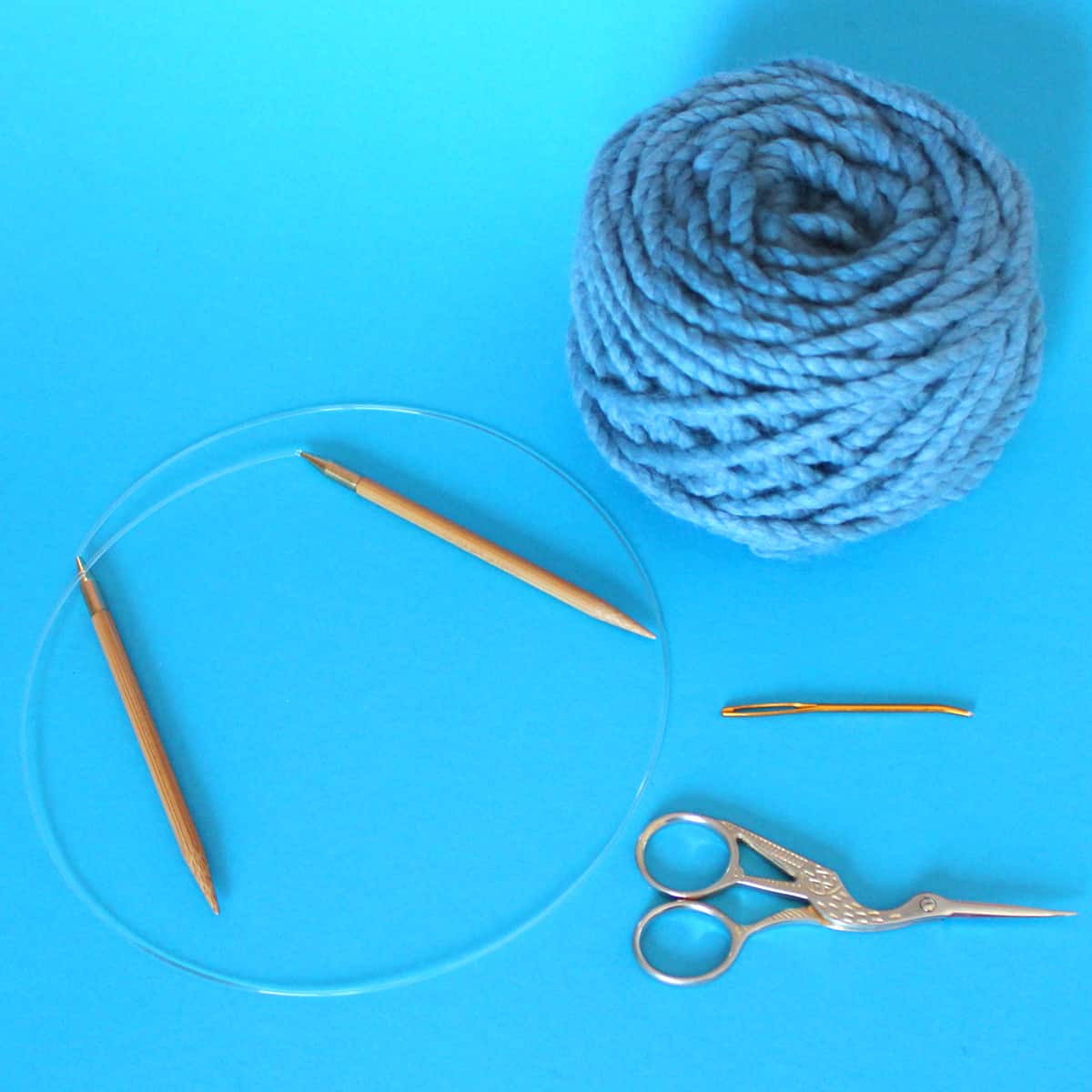 Knitting supplies of circular needle, tapestry needle, scissors, and super bulky yarn in blue color.