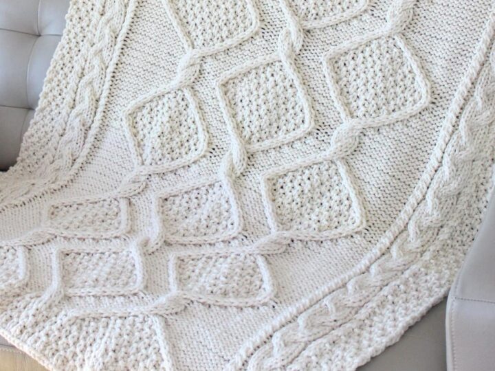 Cable knit blanket in diamond moss stitch draped on a couch in cream colored yarn.
