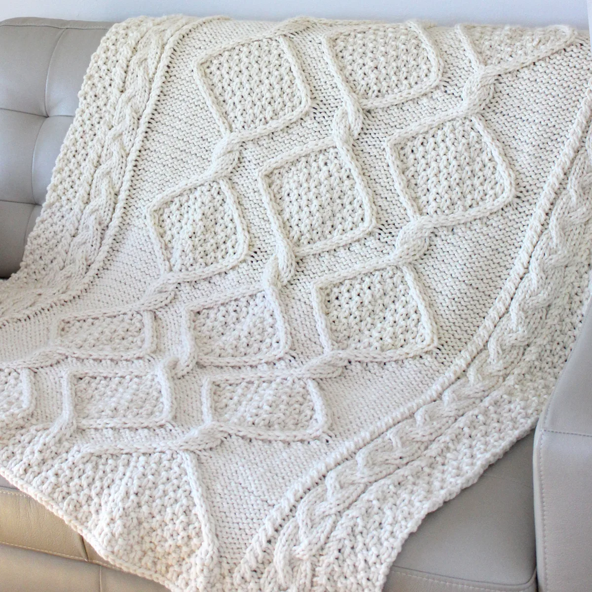 Cable knit blanket in diamond moss stitch draped on a couch in cream colored yarn.