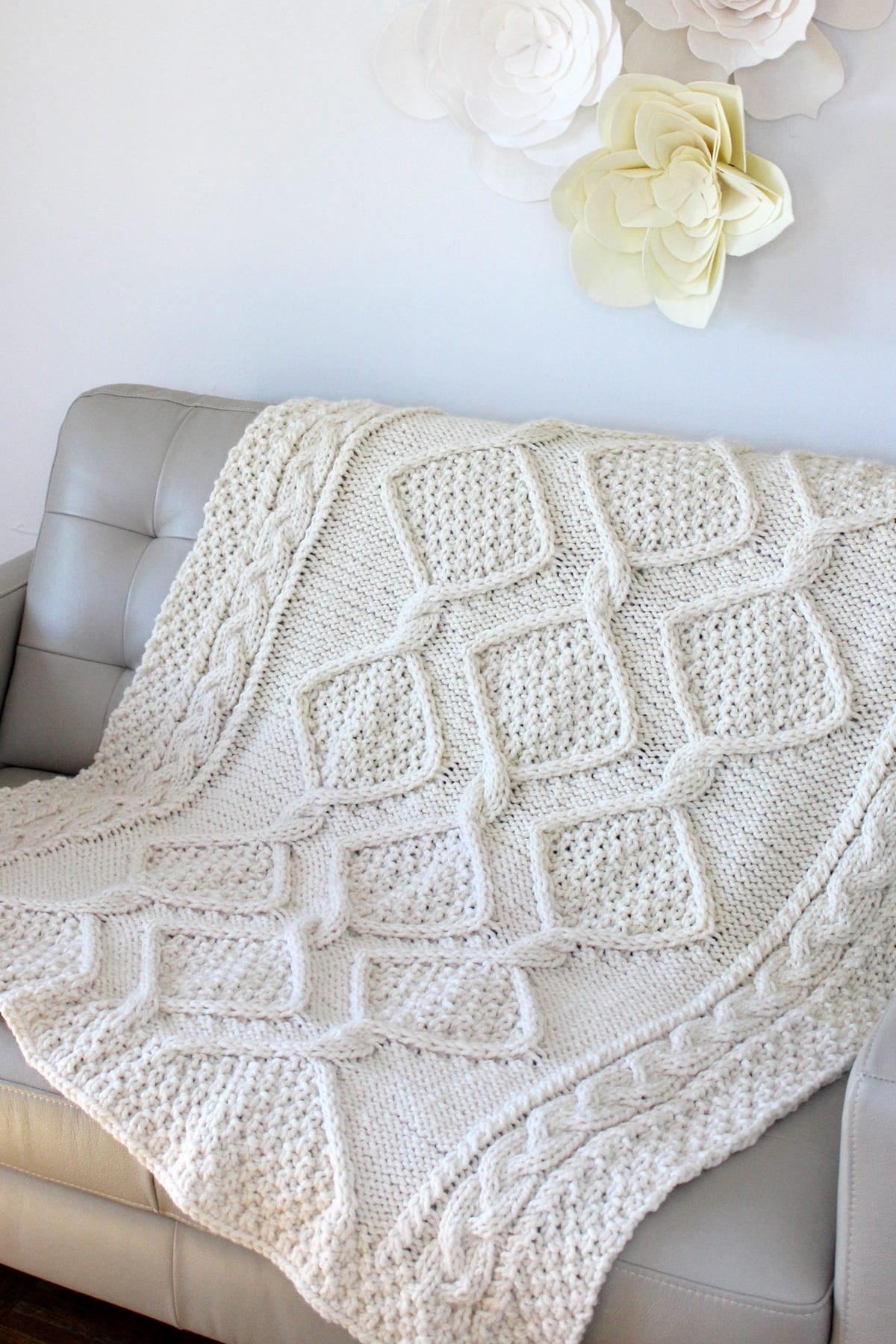 Cable knit blanket draped on a couch in cream colored yarn.
