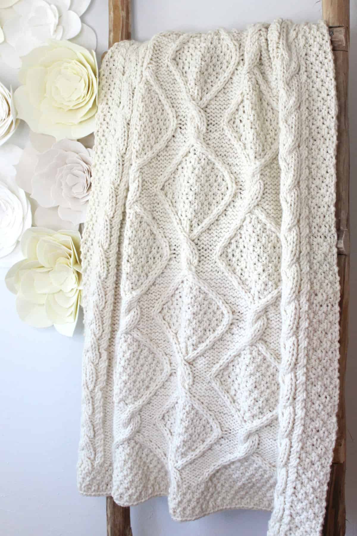 Cable knit blanket throw hung on a wooden ladder in cream colored yarn.