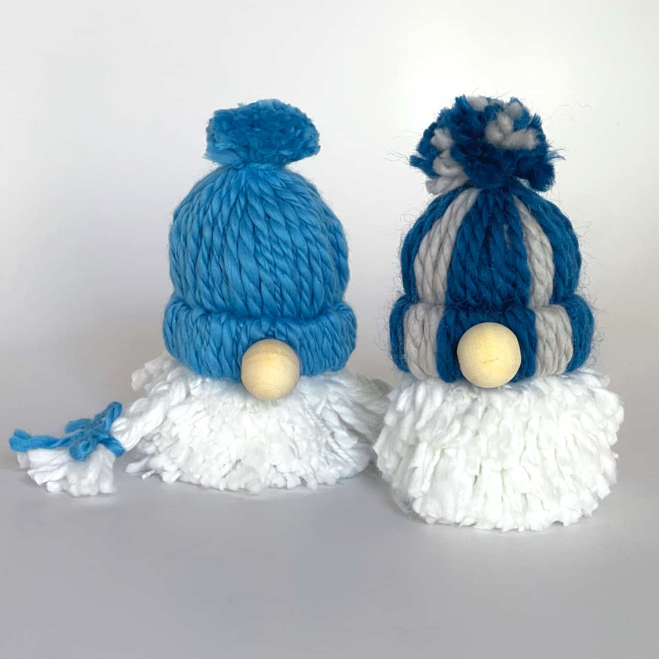 Two yarn gnome ornaments made with pom poms and a wooden nose in blue and white colors.