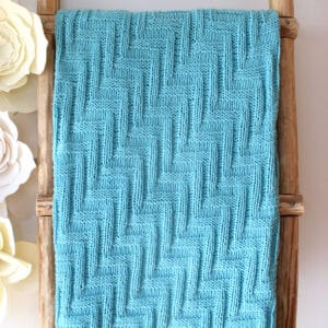 Knitted Blanket in zigzag stitch texture displayed on a wooden ladder in blue yarn color.