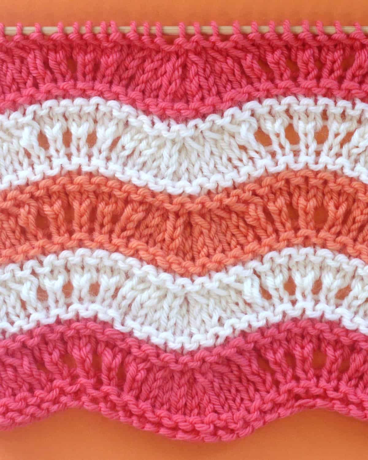 Ripple Ridge knit stitch texture in alternating yarn colors of pink, orange, and white on a wooden bamboo knitting needle.