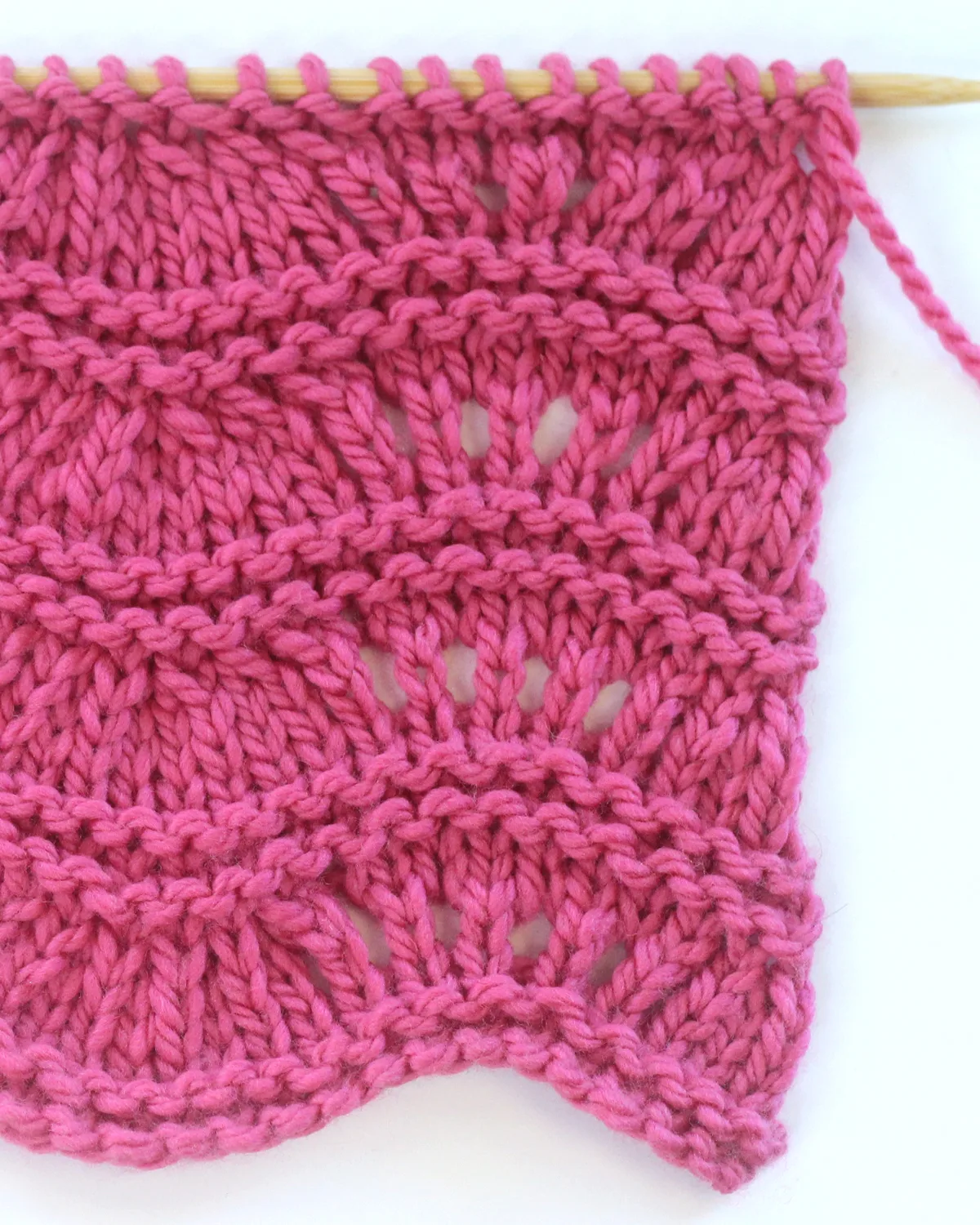 Close up of Ripple Ridge knit stitch texture in bulky weight dark pink colored yarn on a wooden bamboo knitting needle.