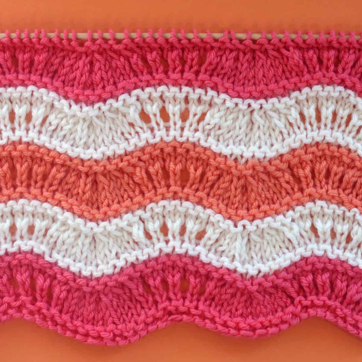 Ripple Ridge knit stitch texture in alternating yarn colors of pink, orange, and white on a wooden bamboo knitting needle.