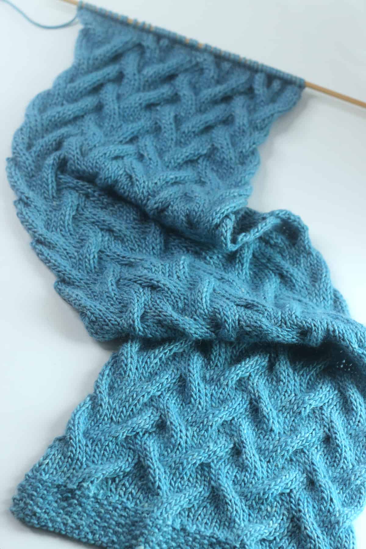 Long scarf in Sand Cable knit stitch pattern knitted flat with a straight knitting needle in blue yarn color.