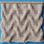Sand Cable stitch on wooden bamboo knitting needle with beige colored yarn on a blue background.