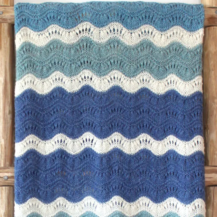 Mavericks Wave Ripple blanket knitted in shades of blue and white displayed on a wooden ladder.