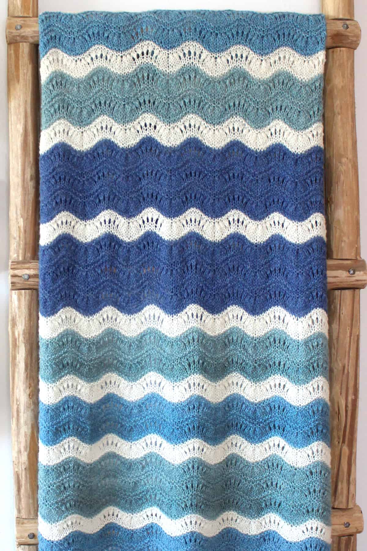 Ripple lace blanket knitted in shades of blue and white displayed on a wooden ladder.