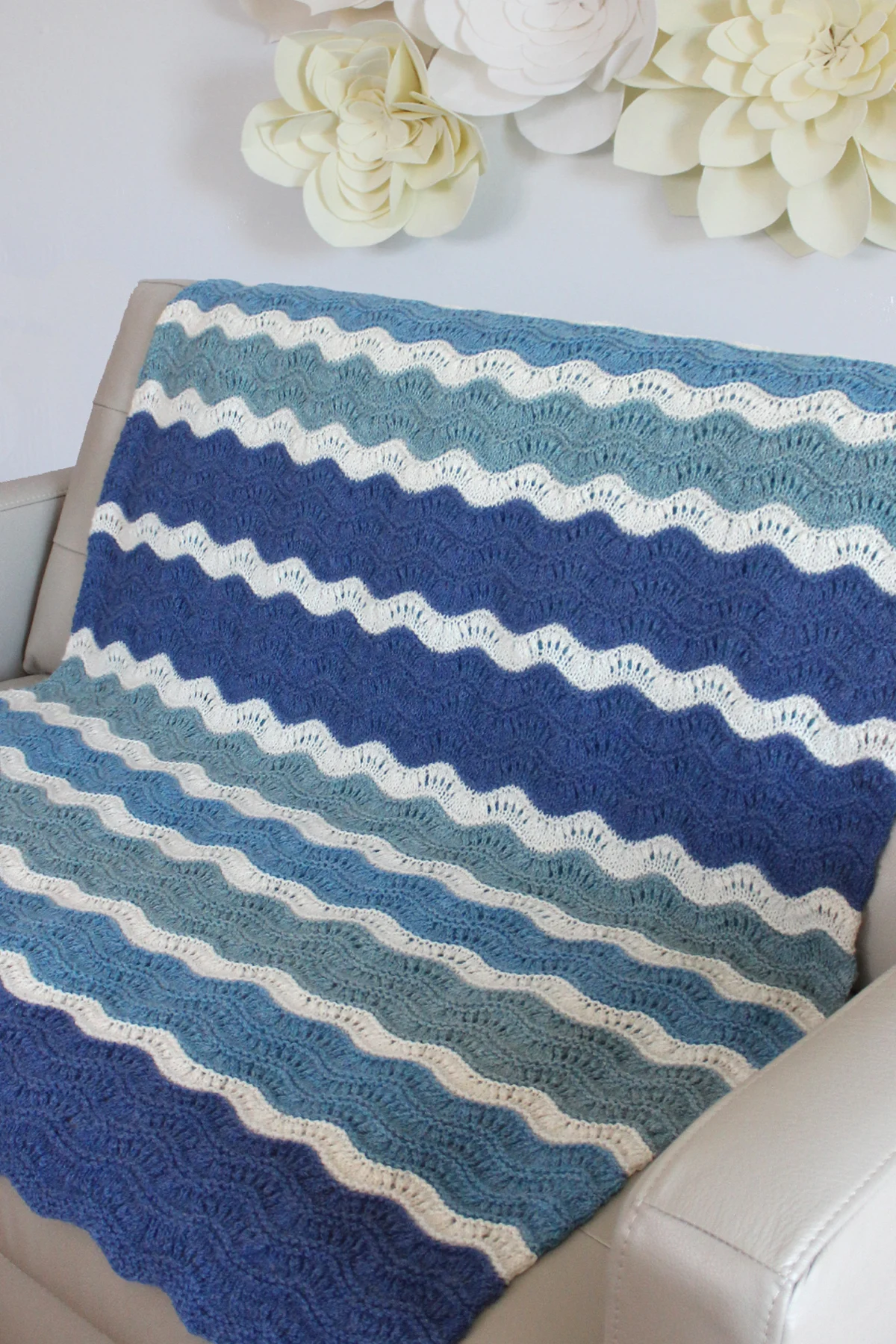 Mavericks Wave Blanket knitted in shades of blue and white displayed on a beige leather couch with white felt flowers on the wall.