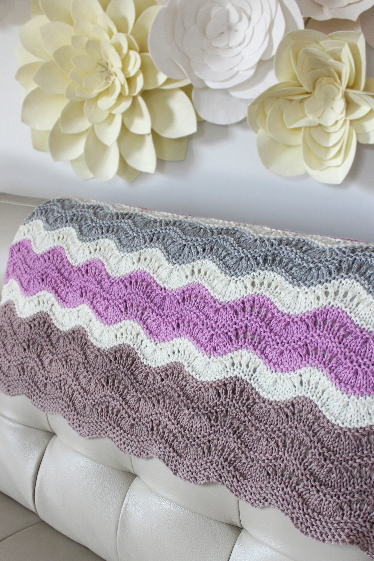 Mavericks Wave Blanket knitted in shades of light purple, brown, grey, and white displayed on a beige leather couch with white felt flowers on the wall.