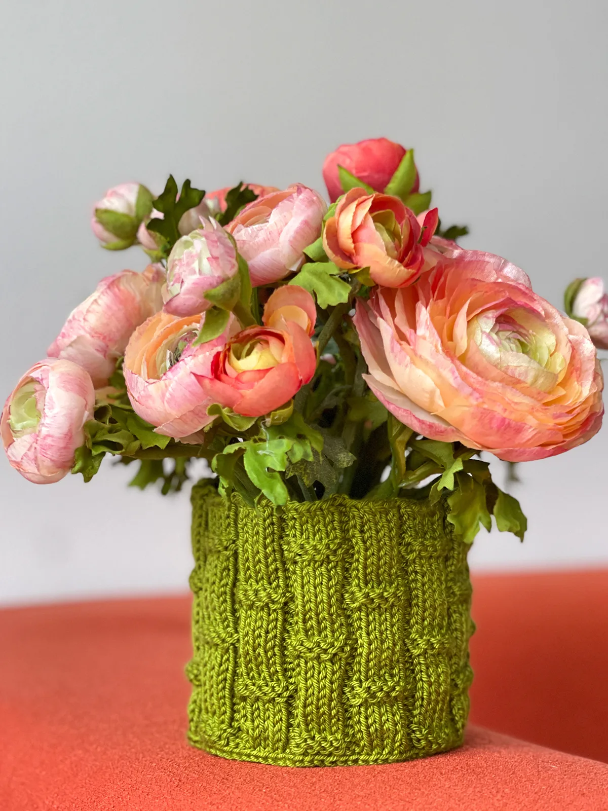 Knitted bamboo textured flower pot cover in green yarn holding peach and pink colored peony flowers.