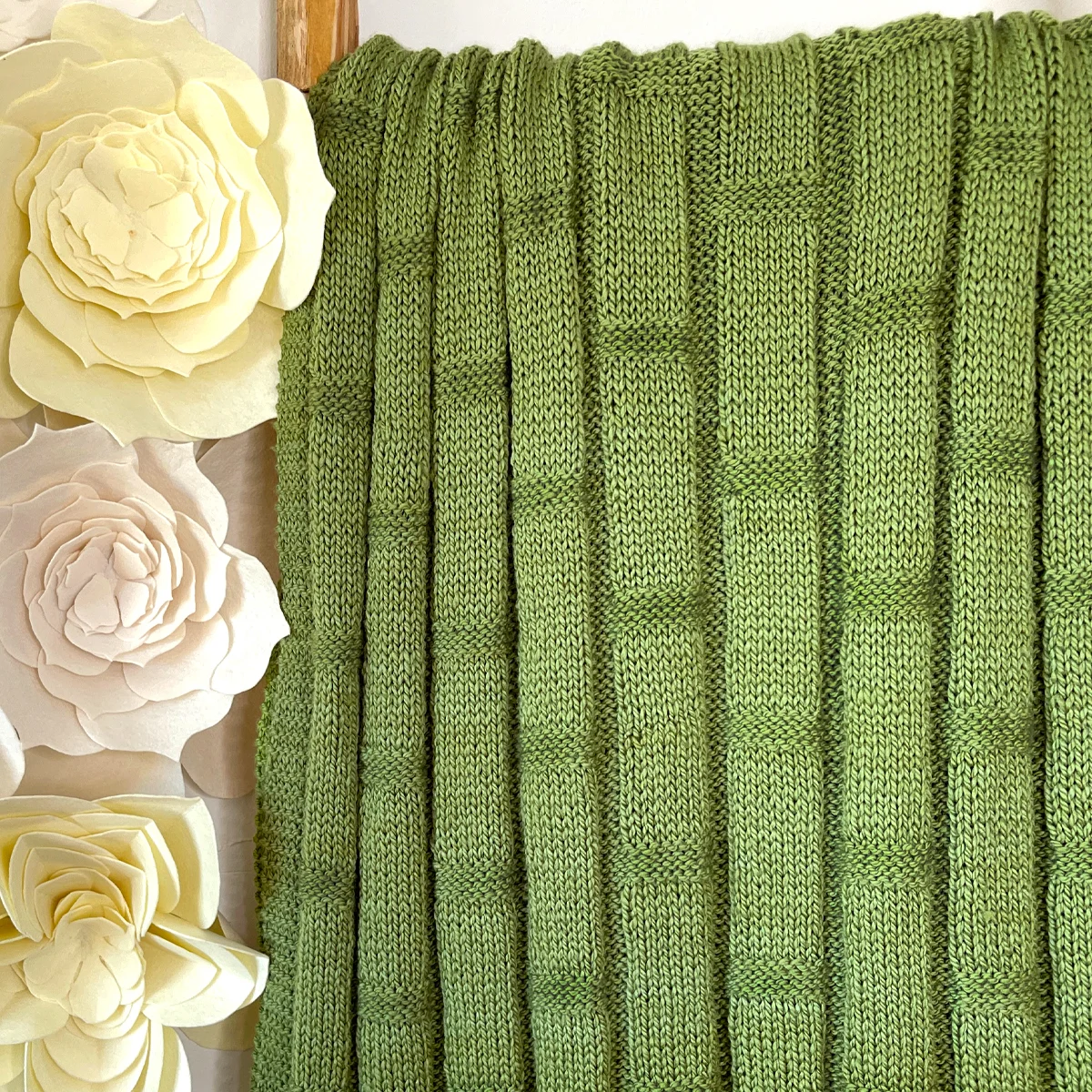 Knitted blanket in bamboo stitch texture in green colored yarn displayed on a wooden ladder.