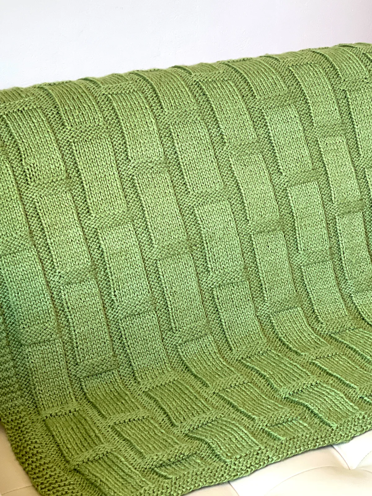 Knitted blanket in bamboo stitch texture in green colored yarn displayed on a loveseat couch.