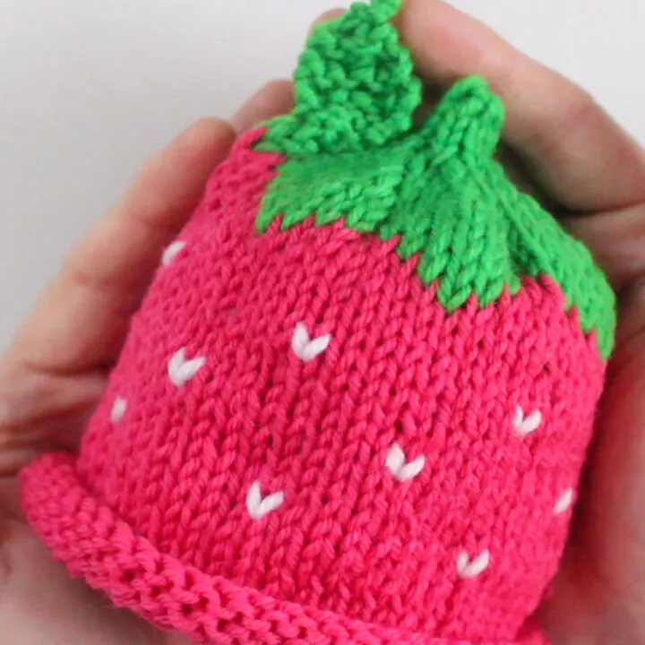 Hands holding a knitted baby hat in strawberry pattern.