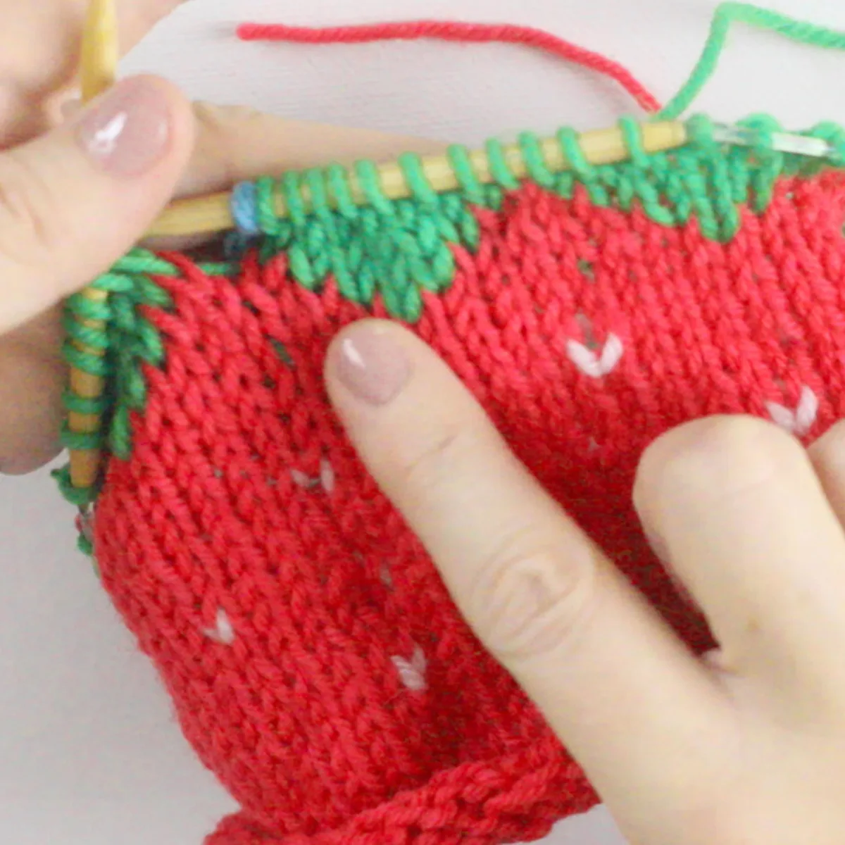 Knitting process of the strawberry baby hat with hands pointing out the topper in green yarn color.
