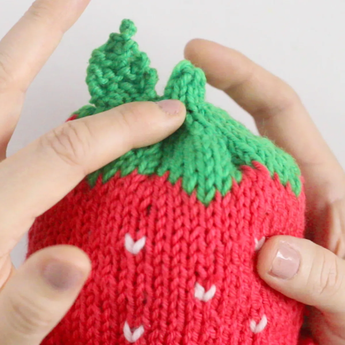 Knitting process of the strawberry baby hat with hands pointing out the top leaf and stem area.