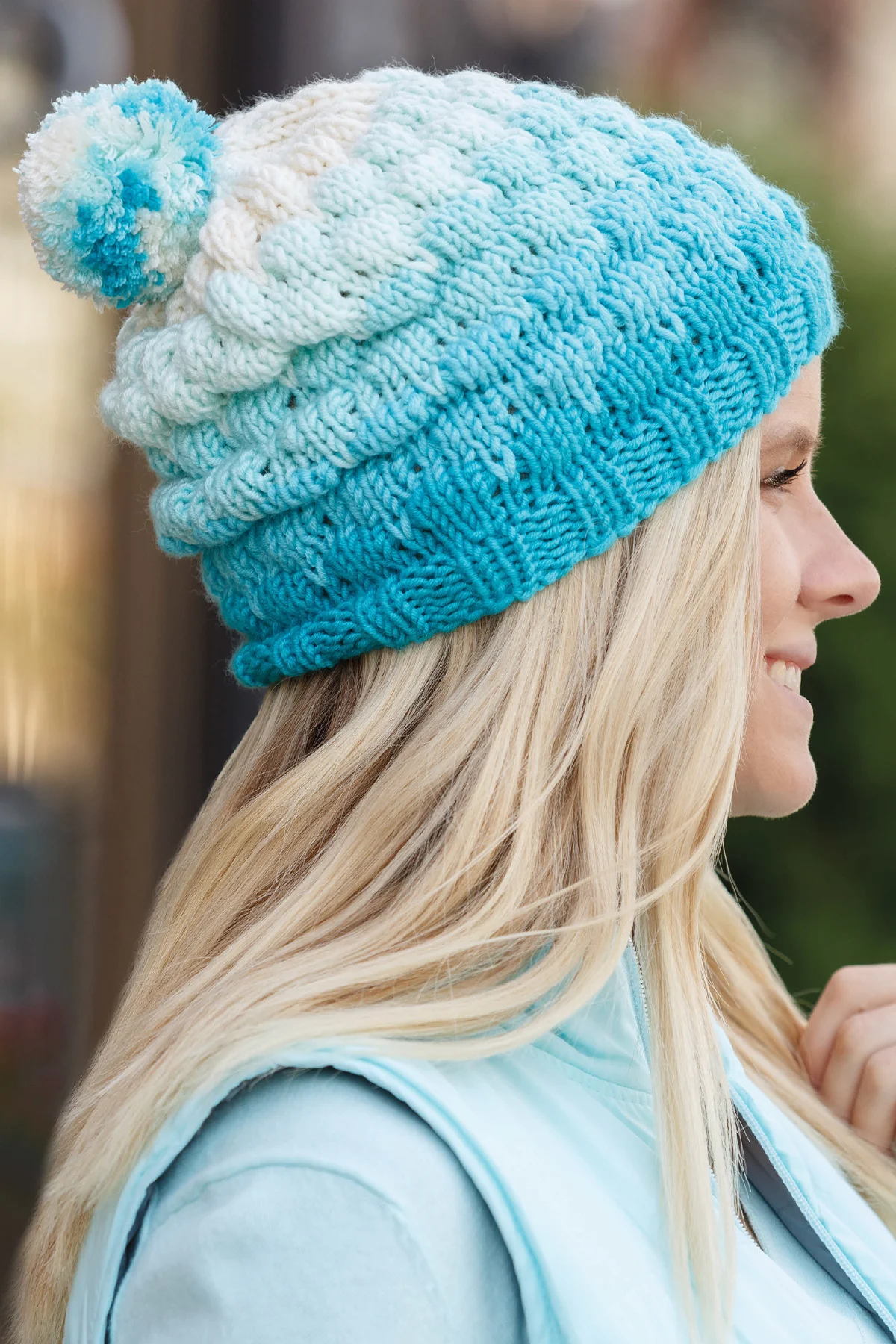Bubble Beanie knitted hat in shades of blue and white yarn colors topped with a pompom worn by a woman with long blonde hair.