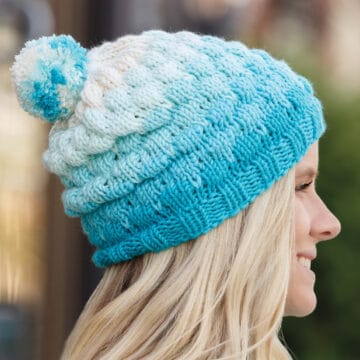 Bubble Beanie knitted hat in shades of blue and white yarn colors topped with a pompom worn by a woman with long blonde hair.