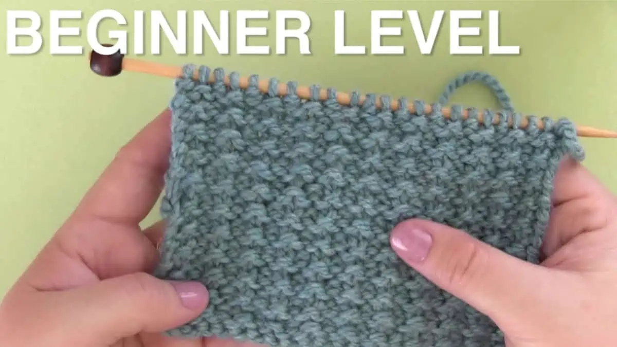 Beginner level with hands holding the Sand Stitch pattern swatch on a knitting needle.
