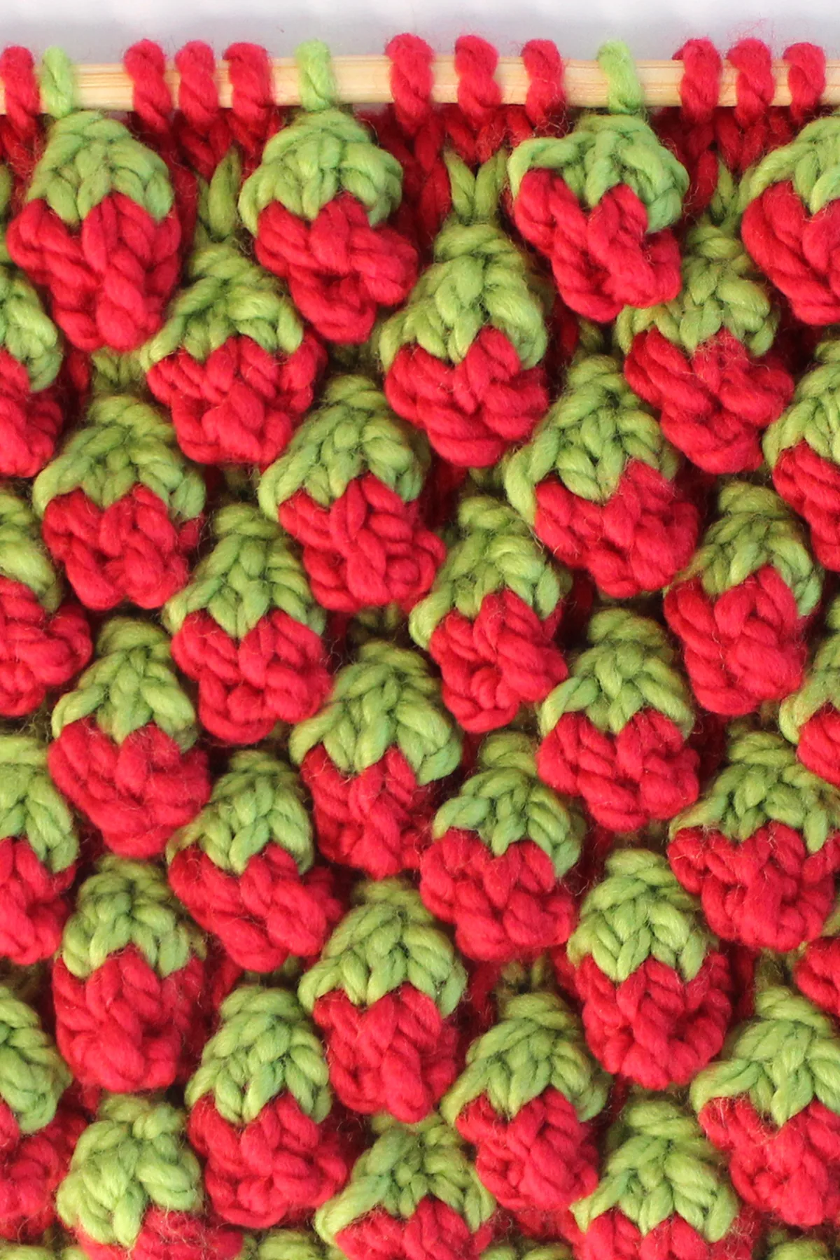 Strawberry knit stitch bobble pattern in red and green colored yarn on a knitting needle.