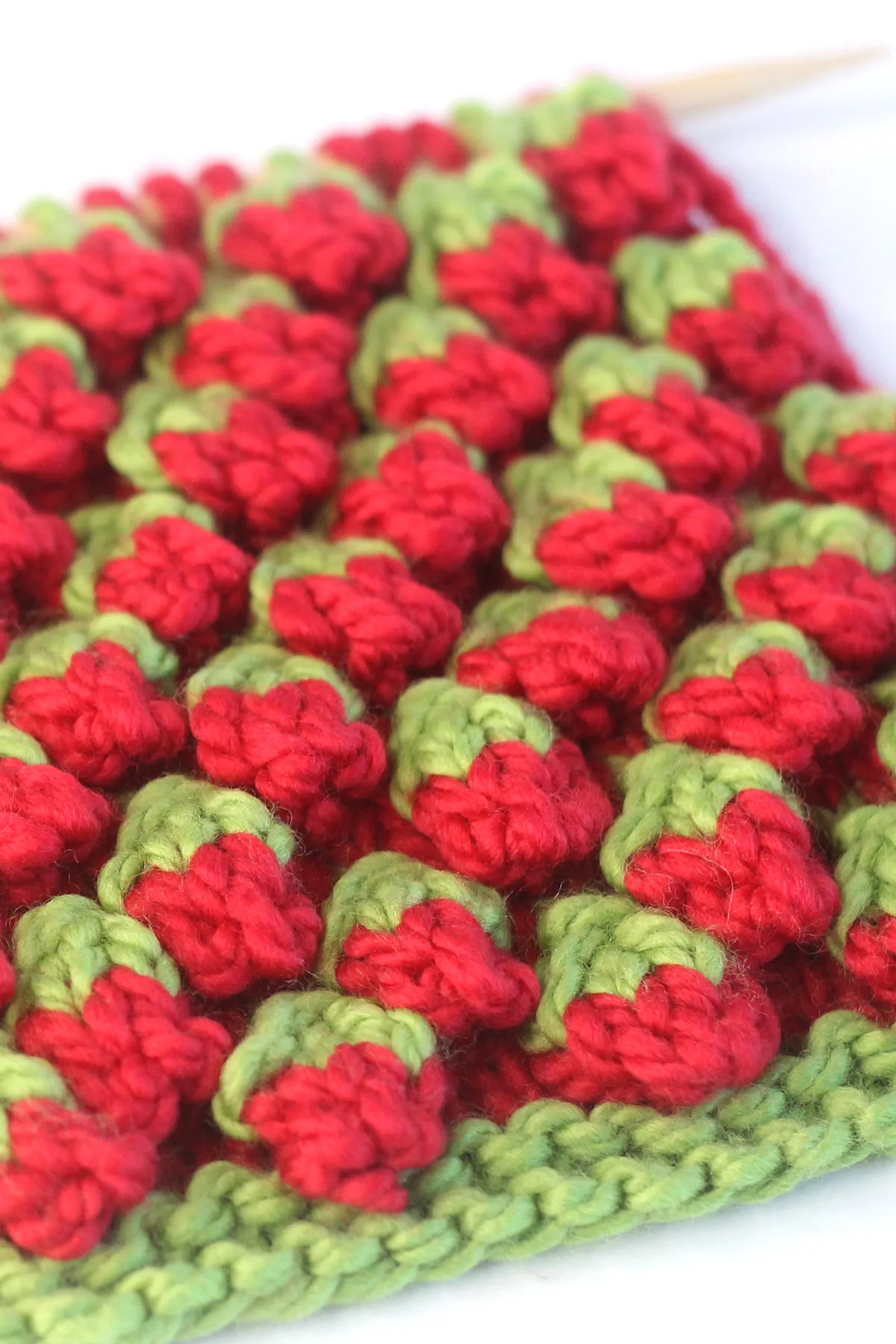 Close-up side view of the strawberry knit stitch bobble pattern in red and green colored yarn on a knitting needle.