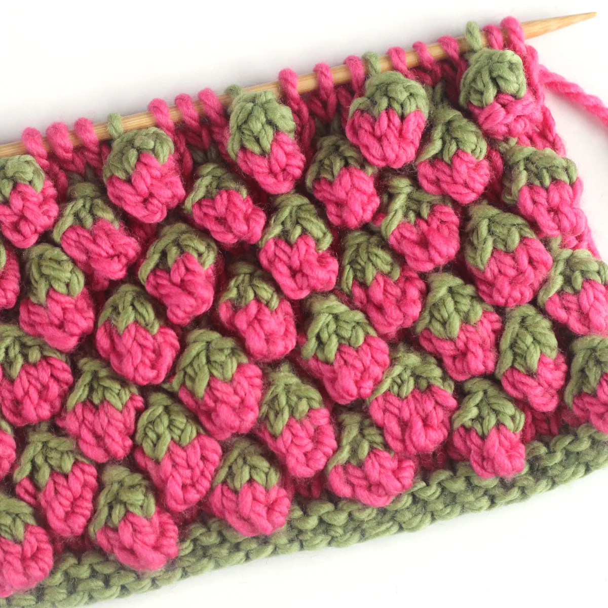 Strawberry knit stitch bobble pattern in pink and green colored yarn on a knitting needle.