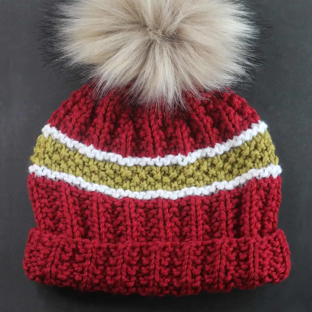 Knitted beanie in red, gold, and white colors with faux fur pom pom.