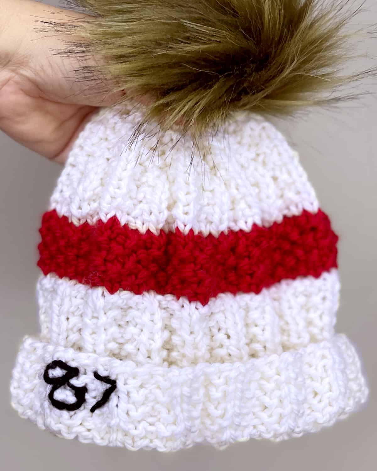 White and red knitted Taylor Swift's 87 Beanie Hat held by a hand.