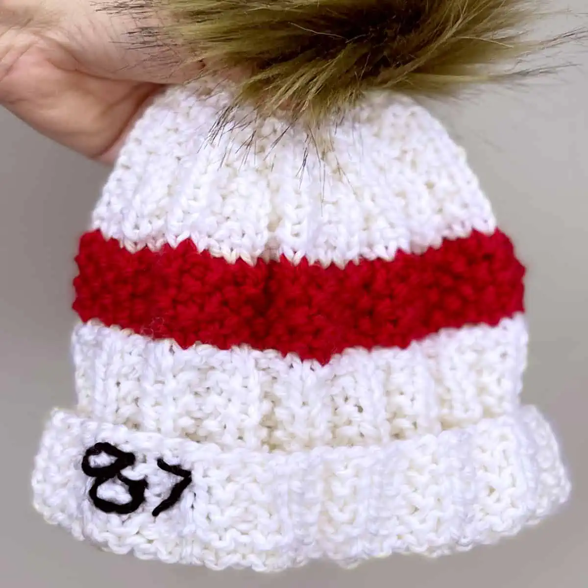 White and red knitted Taylor Swift's 87 Beanie Hat held by a hand.