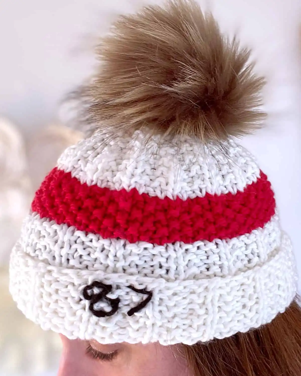 Woman wearing white and red ribbed knit beanie with 87 embroidered and faux fur pom pom.