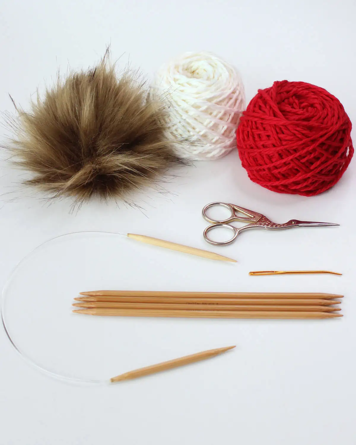 Knitting supplies include faux fur pom pom, red and white yarn, scissors, circular needle, double-pointed needles, and a tapestry needle.
