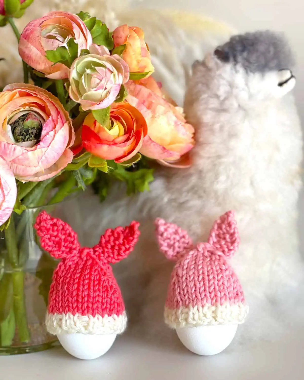 Knitted Bunny Egg Cozies in peach and pink yarn colors beside a llama toy and flower vase.